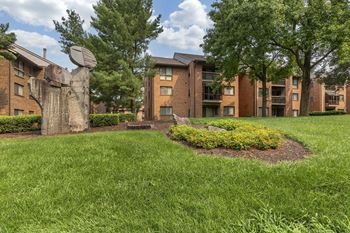 A grassy area in front of apartment buildings with a sculpture and trees.
